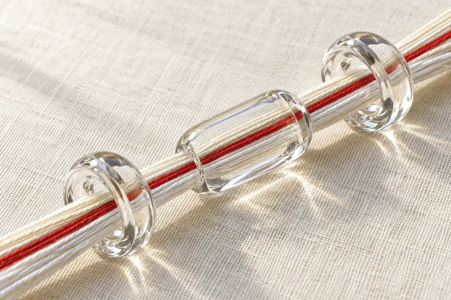 Un faisceau de fils blancs et d’un fil rouge passe dans trois guides en verre posés sur du lin.